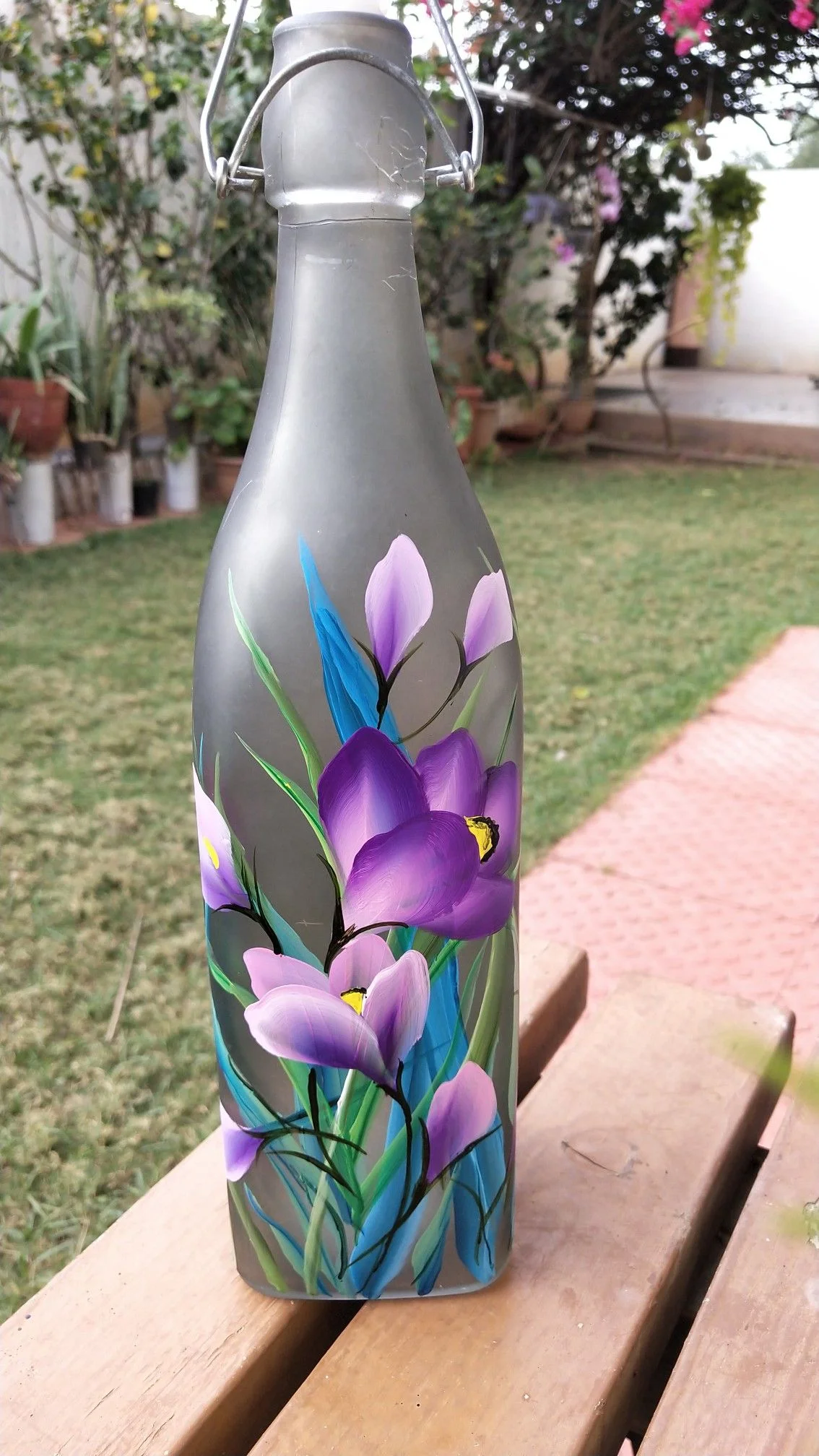 Chai thủy tinh màu bạc vẽ hoa sen tím đặt trên ghế gỗ ngoài vườn